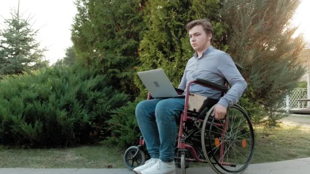 Een gehandicapte man in een rolstoel zit en werken op een laptop in het park — Stockvideo