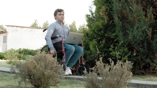 Un hombre discapacitado está sentado en una silla de ruedas y trabajando en un portátil en el parque — Vídeo de stock