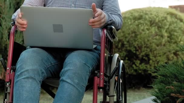 Un disabile è seduto su una sedia a rotelle e lavora su un computer portatile nel parco — Video Stock