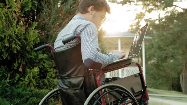Niepełnosprawny mężczyzna siedzi na wózku inwalidzkim i działa na laptopie w parku — Wideo stockowe