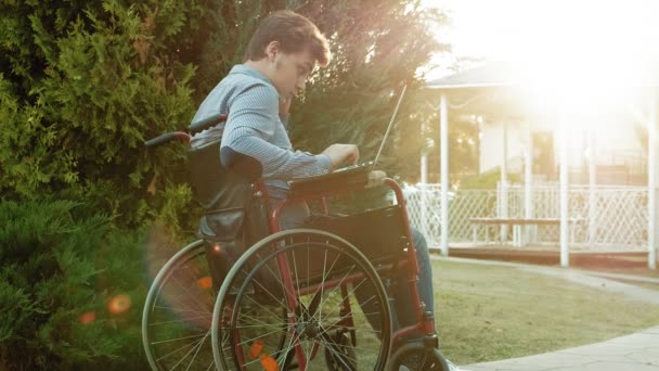 Un handicapé est assis dans un fauteuil roulant et travaille sur un ordinateur portable dans le parc — Video