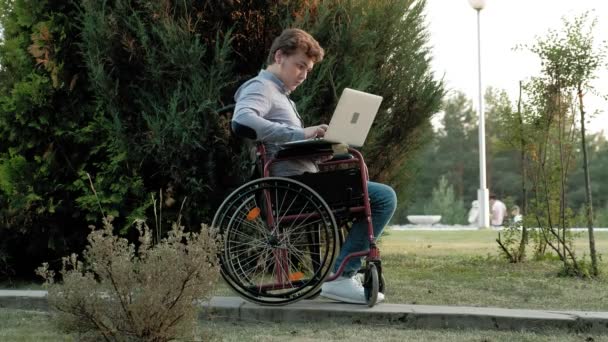 Um deficiente está sentado em uma cadeira de rodas e trabalhando em um laptop no parque — Vídeo de Stock