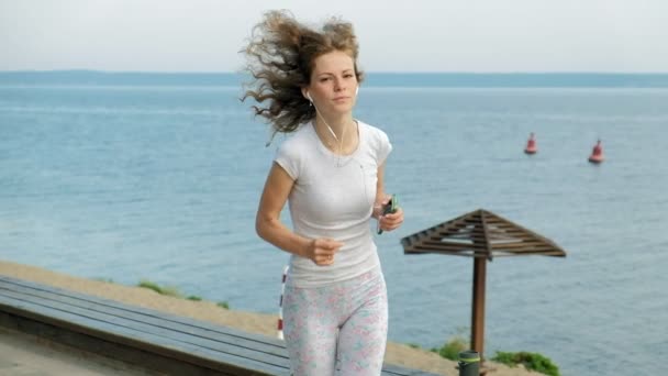 Une jeune femme avec une belle silhouette est engagée dans la gymnastique dans la mer à l'aube. Elle court le long de la côte dans les écouteurs, Super slow motion — Video