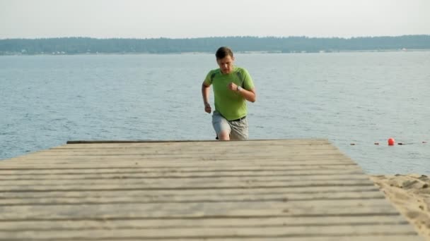 Dojrzały człowiek jest zaangażowany w gimnastyce w morzu o świcie. Ona biegnie wzdłuż wybrzeża w słuchawkach, Super slow motion — Wideo stockowe