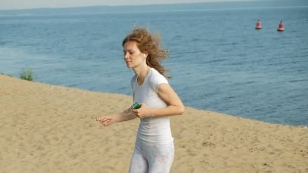 Une jeune femme avec une belle silhouette est engagée dans la gymnastique dans la mer à l'aube. Elle court le long de la côte dans les écouteurs, Super slow motion — Video