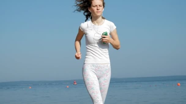 Eine junge Frau mit schöner Figur turnt im Morgengrauen im Meer. sie rennt mit Kopfhörern an der Küste entlang, Super-Zeitlupe — Stockvideo