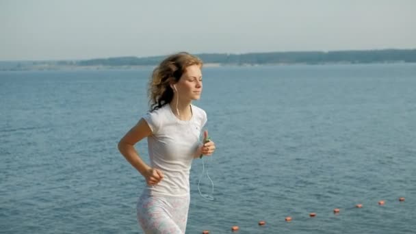 Uma jovem mulher com uma boa figura está envolvida em ginástica no mar ao amanhecer. Ela corre ao longo do litoral em fones de ouvido, Super câmera lenta — Vídeo de Stock