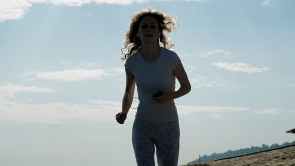 Une jeune femme avec une belle silhouette est engagée dans la gymnastique dans la mer à l'aube. Elle court le long de la côte dans les écouteurs, Super slow motion — Video