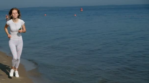 Uma jovem mulher com uma boa figura está envolvida em ginástica no mar ao amanhecer. Ela corre ao longo do litoral em fones de ouvido, Super câmera lenta — Vídeo de Stock