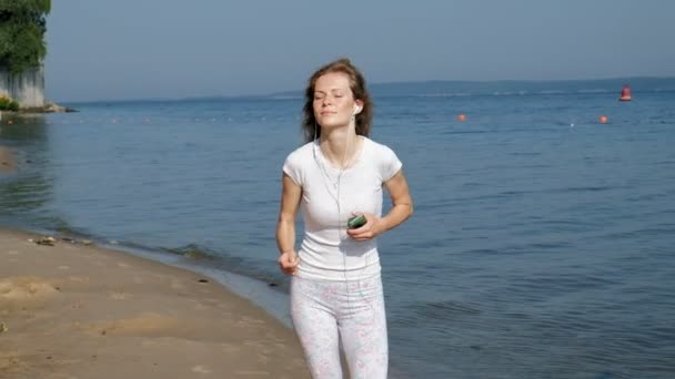 Una joven con una bella figura se dedica a la gimnasia en el mar al amanecer. Ella corre a lo largo de la costa en auriculares, Super cámara lenta — Vídeo de stock