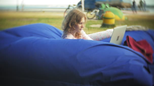 Young woman uses a laptop, lying in a lounge area of the park on soft puffs — Stock Video