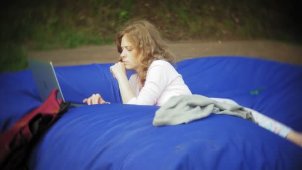 Νεαρή γυναίκα χρησιμοποιεί το φορητό υπολογιστή, ξαπλωμένη στο σαλόνι του πάρκου με μαλακό εισπνοές — Αρχείο Βίντεο