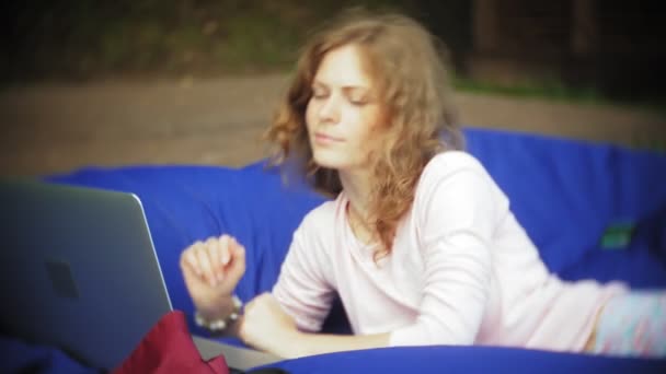 Mujer joven utiliza un ordenador portátil, acostado en una sala de estar del parque en soplos suaves — Vídeo de stock