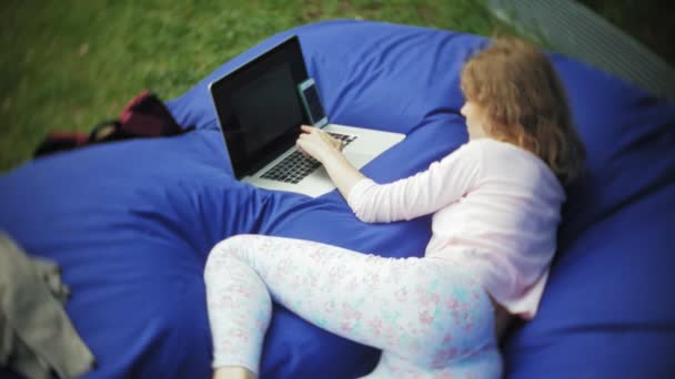 Mujer joven utiliza un ordenador portátil, acostado en una sala de estar del parque en soplos suaves — Vídeos de Stock
