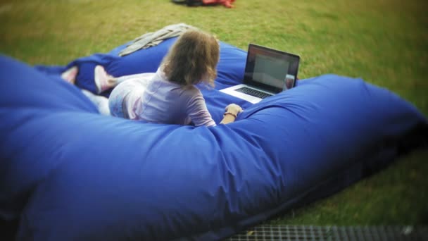 Jeune femme utilise un ordinateur portable, couché dans un coin salon du parc sur des bouffées molles — Video