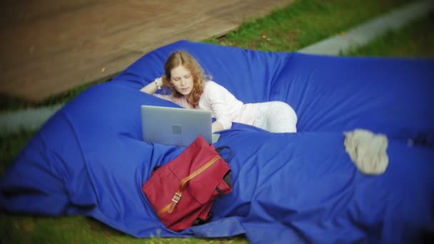 若い女性は、柔らかなパフでパークのラウンジ エリアで横になっているラップトップを使用してください。 — ストック動画