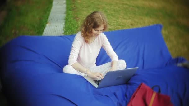 Genç kadın bir dizüstü bilgisayar, yumuşak puffs üzerinde Park lounge alanında yalan kullanır — Stok video