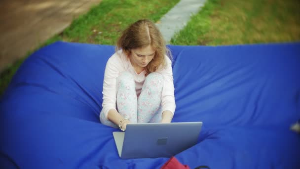 Junge Frau benutzt einen Laptop, der in einem Lounge-Bereich des Parks auf weichen Windbeutel liegt — Stockvideo