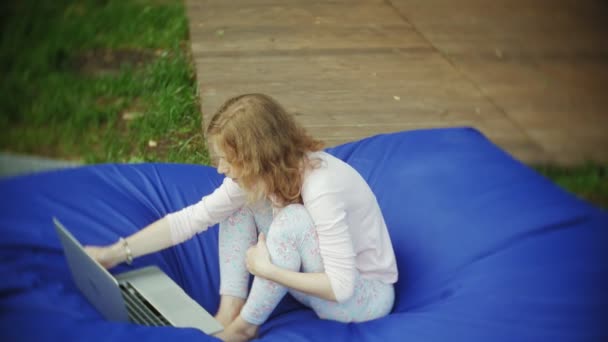 Молода жінка використовує ноутбук, лежачи в лаунж-зоні парку на м'яких пуфах — стокове відео