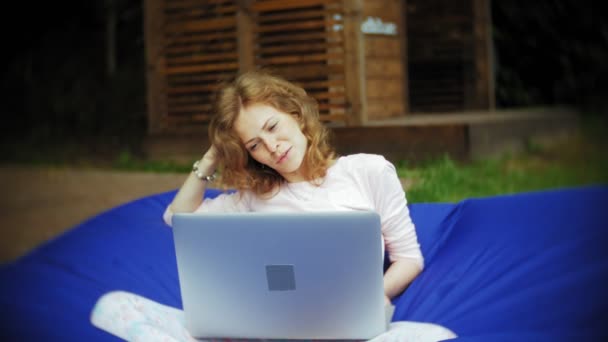 Νεαρή γυναίκα χρησιμοποιεί το φορητό υπολογιστή, ξαπλωμένη στο σαλόνι του πάρκου με μαλακό εισπνοές — Αρχείο Βίντεο