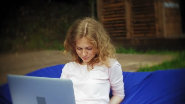 Junge Frau benutzt einen Laptop, der in einem Lounge-Bereich des Parks auf weichen Windbeutel liegt — Stockvideo