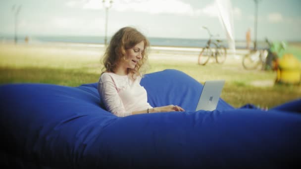 युवा महिला एक लैपटॉप का उपयोग करती है, नरम पफ पर पार्क के एक लाउंज क्षेत्र में पड़ी है — स्टॉक वीडियो