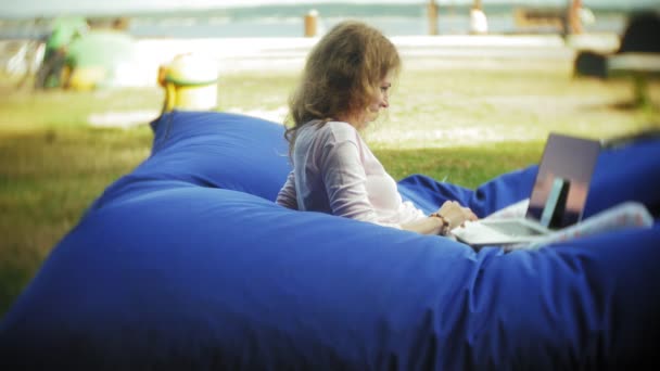 Jonge vrouw maakt gebruik van een laptop, liggend in een lounge van het park op zachte soezen — Stockvideo