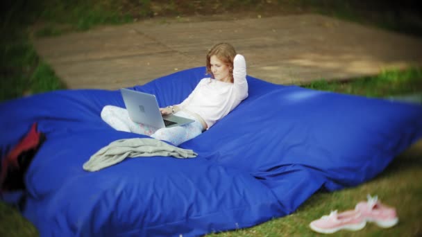 Jonge vrouw maakt gebruik van een laptop, liggend in een lounge van het park op zachte soezen — Stockvideo