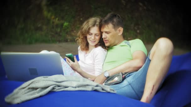 Volwassen man en jonge vrouw gebruikt een laptop, liggend in een lounge van het park op zachte soezen — Stockvideo