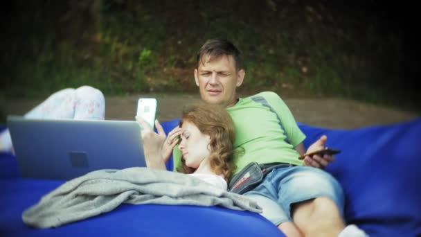 Молода жінка і зрілий чоловік використовує ноутбук, лежачи в лаунж-зоні парку на м'яких пуфах — стокове відео