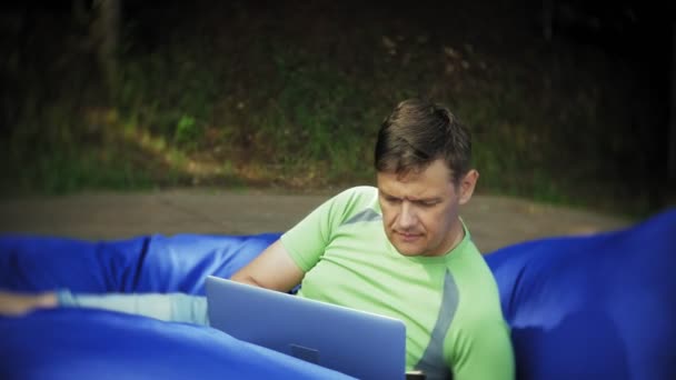 Зрелый мужчина использует ноутбук, лежащий в зоне отдыха парка на мягких слоеных подушечках — стоковое видео