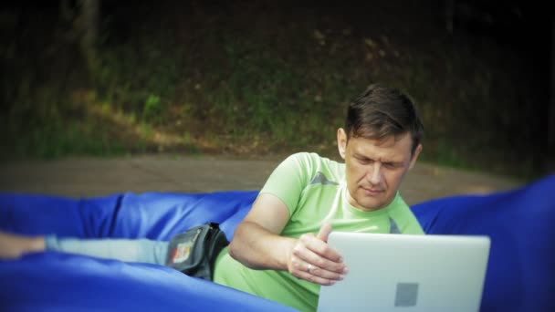 Homme mûr utilise un ordinateur portable, couché dans un coin salon du parc sur des bouffées molles — Video