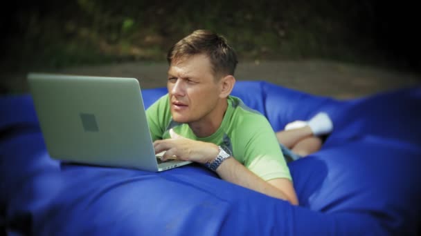 Mature man uses a laptop, lying in a lounge area of the park on soft puffs — Stock Video