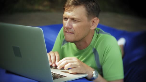 Ein älterer Mann benutzt einen Laptop, der in einem Aufenthaltsraum des Parks auf weichen Windbeutel liegt — Stockvideo