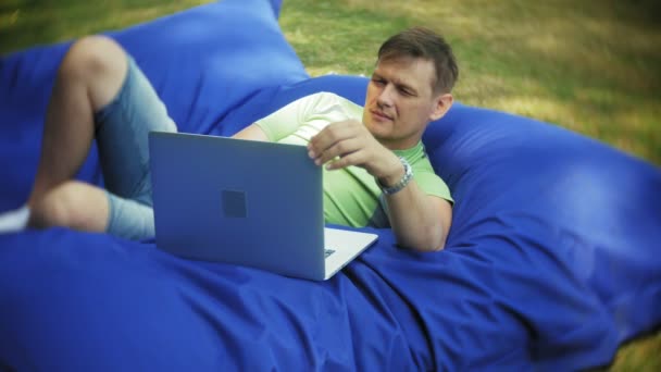 Hombre maduro utiliza un ordenador portátil, acostado en una sala de estar del parque en soplos suaves — Vídeo de stock