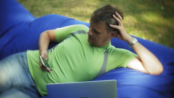 Зрелый мужчина использует ноутбук, лежащий в зоне отдыха парка на мягких слоеных подушечках — стоковое видео