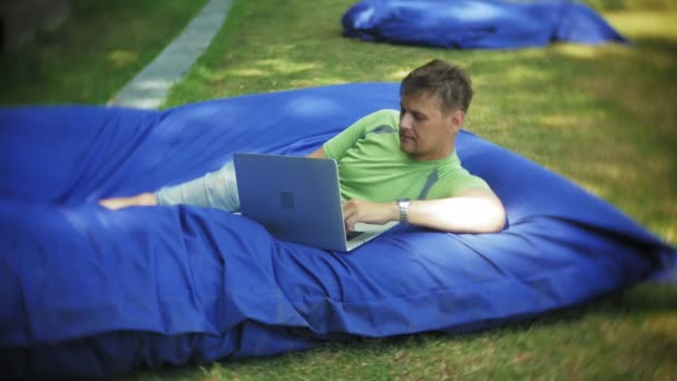 Mature man uses a laptop, lying in a lounge area of the park on soft puffs — Stock Video