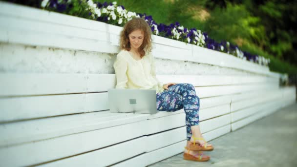 Schöne Frau arbeitet an einem Laptop auf einer Holzbank im Park — Stockvideo