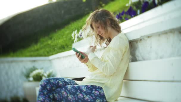 Belle femme utilise un smartphone sur un banc en bois dans le parc — Video