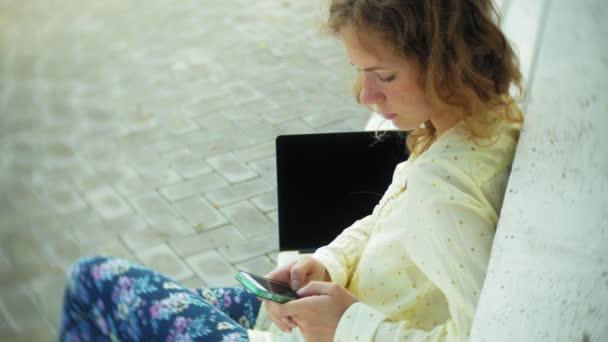 Hermosa mujer utiliza un teléfono inteligente en un banco de madera en el parque — Vídeos de Stock