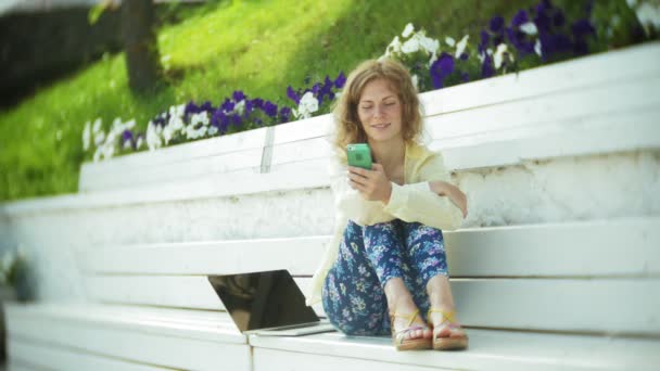 Mulher bonita usa um smartphone em um banco de madeira no parque — Vídeo de Stock