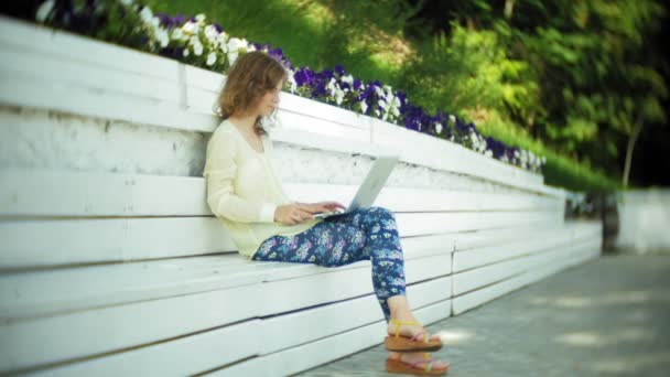 Beautiful woman working on a laptop on a wooden bench in the park — Stock Video