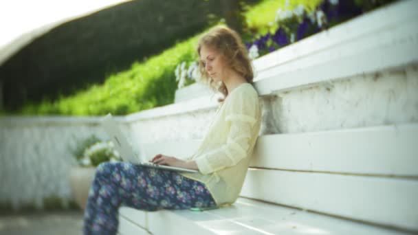 Belle femme travaillant sur un ordinateur portable sur un banc en bois dans le parc — Video