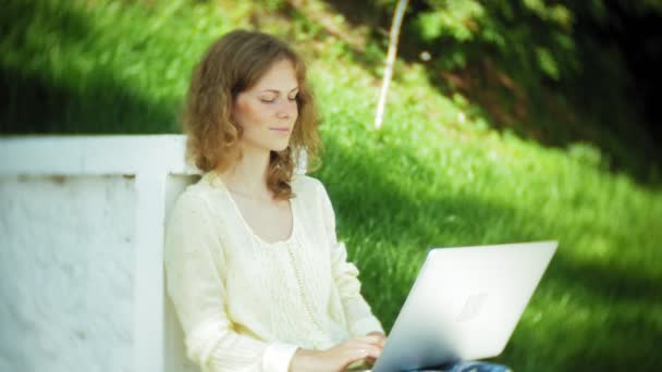 Красива жінка працює на ноутбуці на дерев'яній лавці в парку — стокове відео
