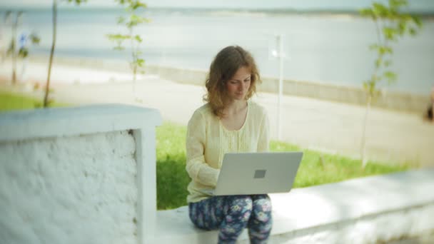 Vacker kvinna arbetar på en bärbar dator på en träbänk i parken — Stockvideo