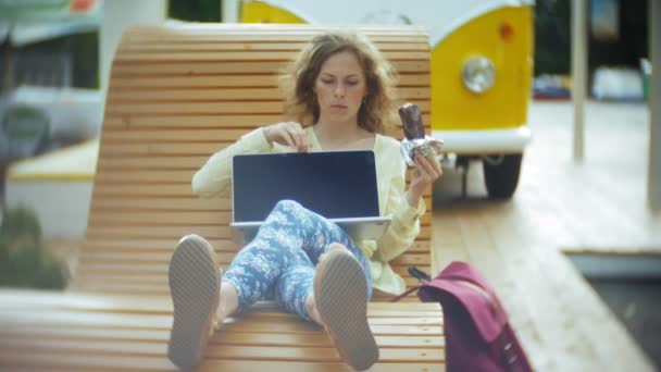 Mulher bonita come um sorvete e trabalha em um laptop em um banco de madeira no parque — Vídeo de Stock