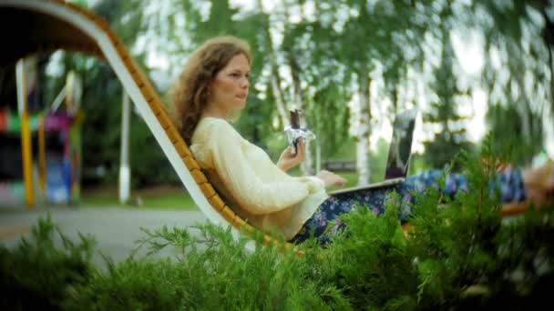 Belle femme mange une crème glacée et travaille sur un ordinateur portable sur un banc en bois dans le parc — Video