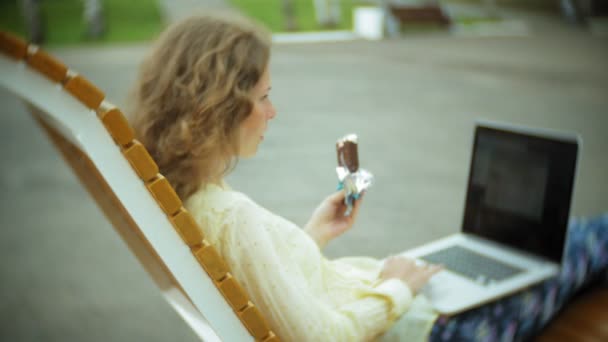Красивая женщина ест мороженое и работает на ноутбуке на деревянной скамейке в парке — стоковое видео