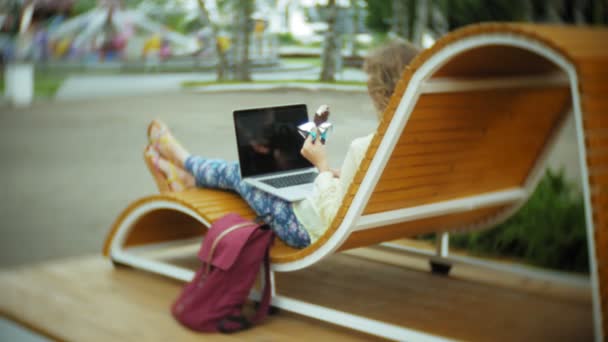 Schöne Frau isst ein Eis und arbeitet an einem Laptop auf einer Holzbank im Park — Stockvideo
