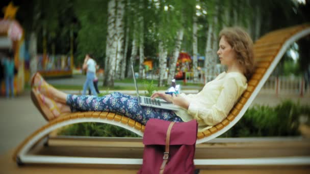 Красива жінка їсть морозиво і працює на ноутбуці на дерев'яній лавці в парку — стокове відео